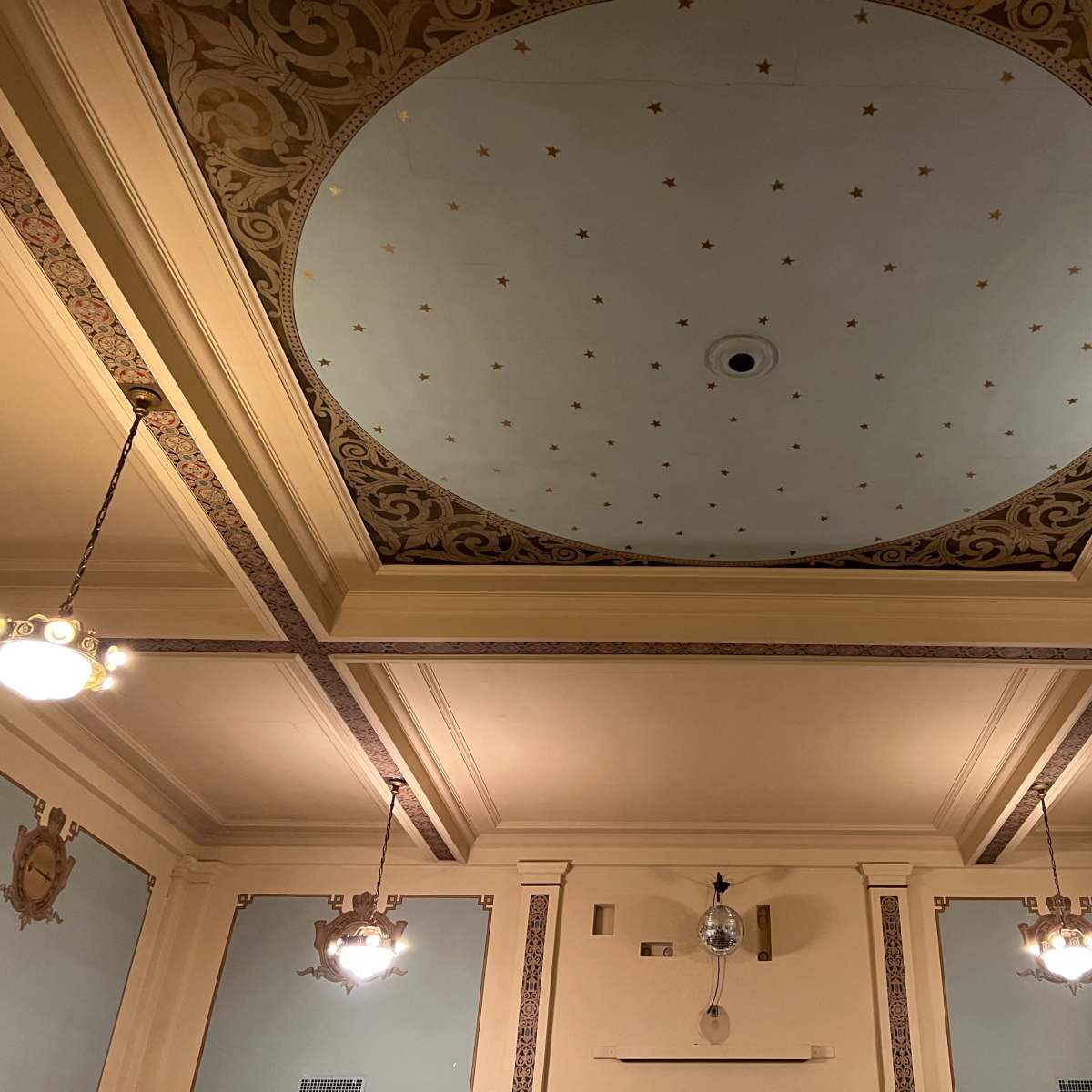 An elaborately decorated ceiling with a central circular design, surrounded by ornate molding and flanked by ornate light fixtures.