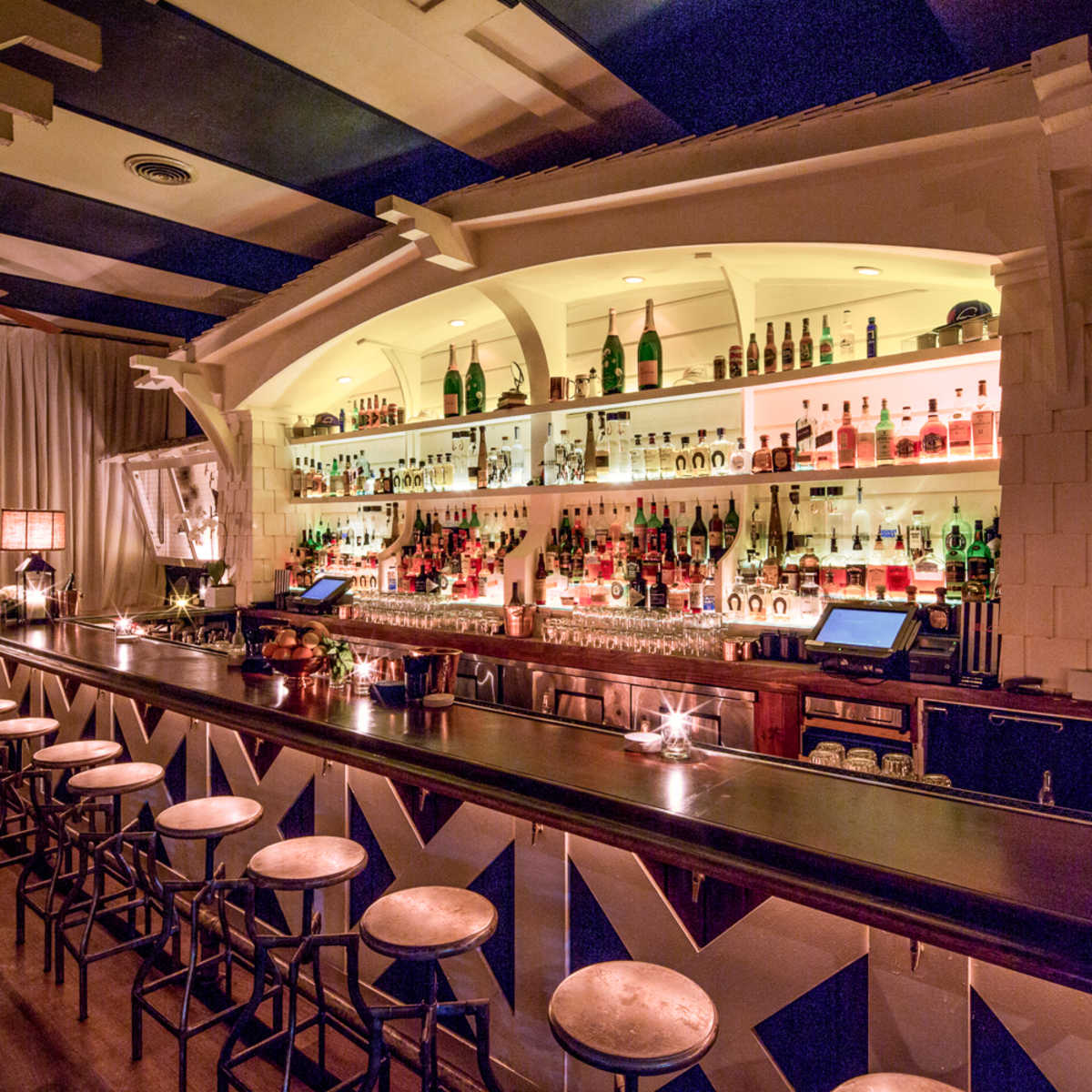 A well-lit bar with a wide selection of bottles displayed on shelves behind the counter and several stools in front.