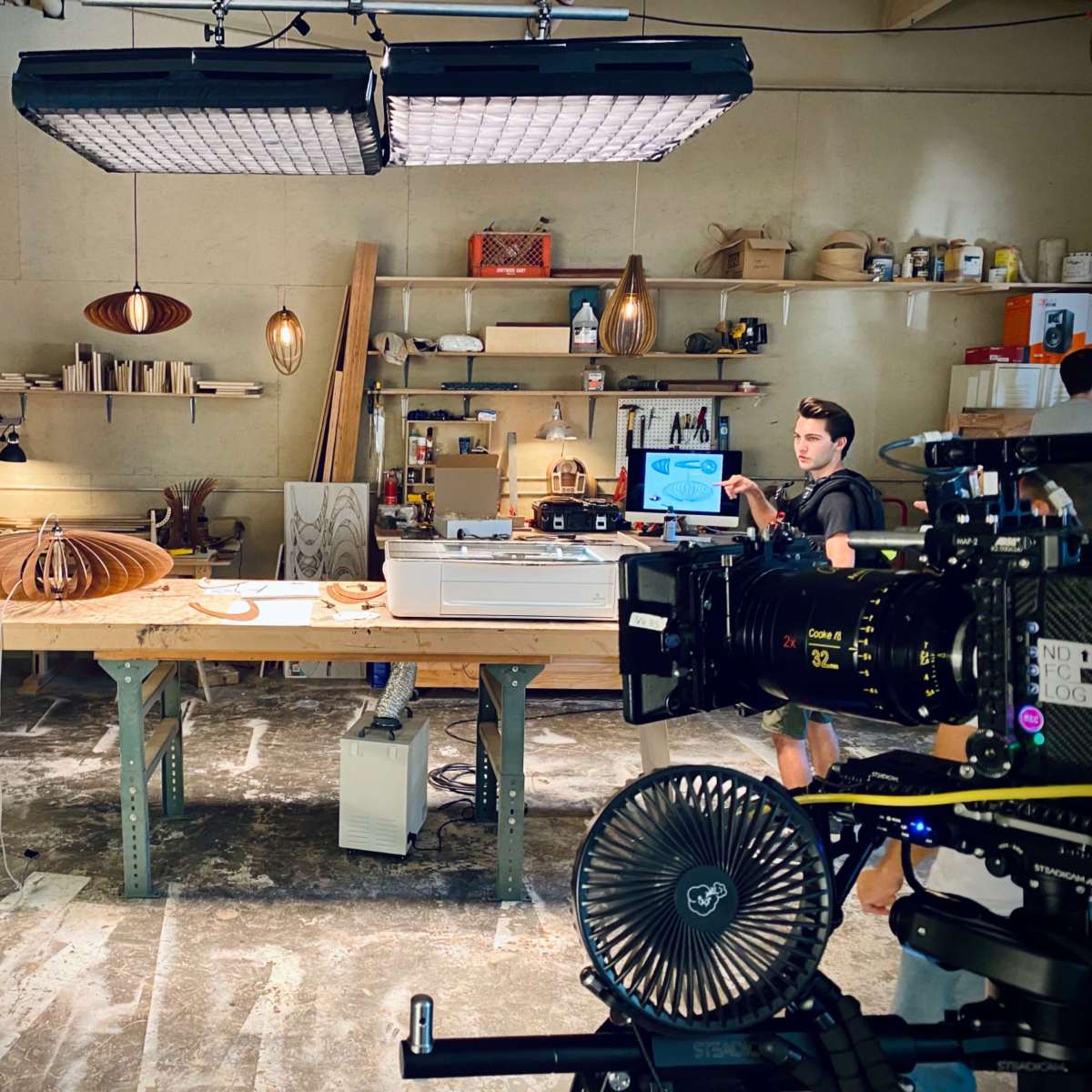 A production set with two men discussing a woodworking project in a workshop equipped with various tools and lighting.