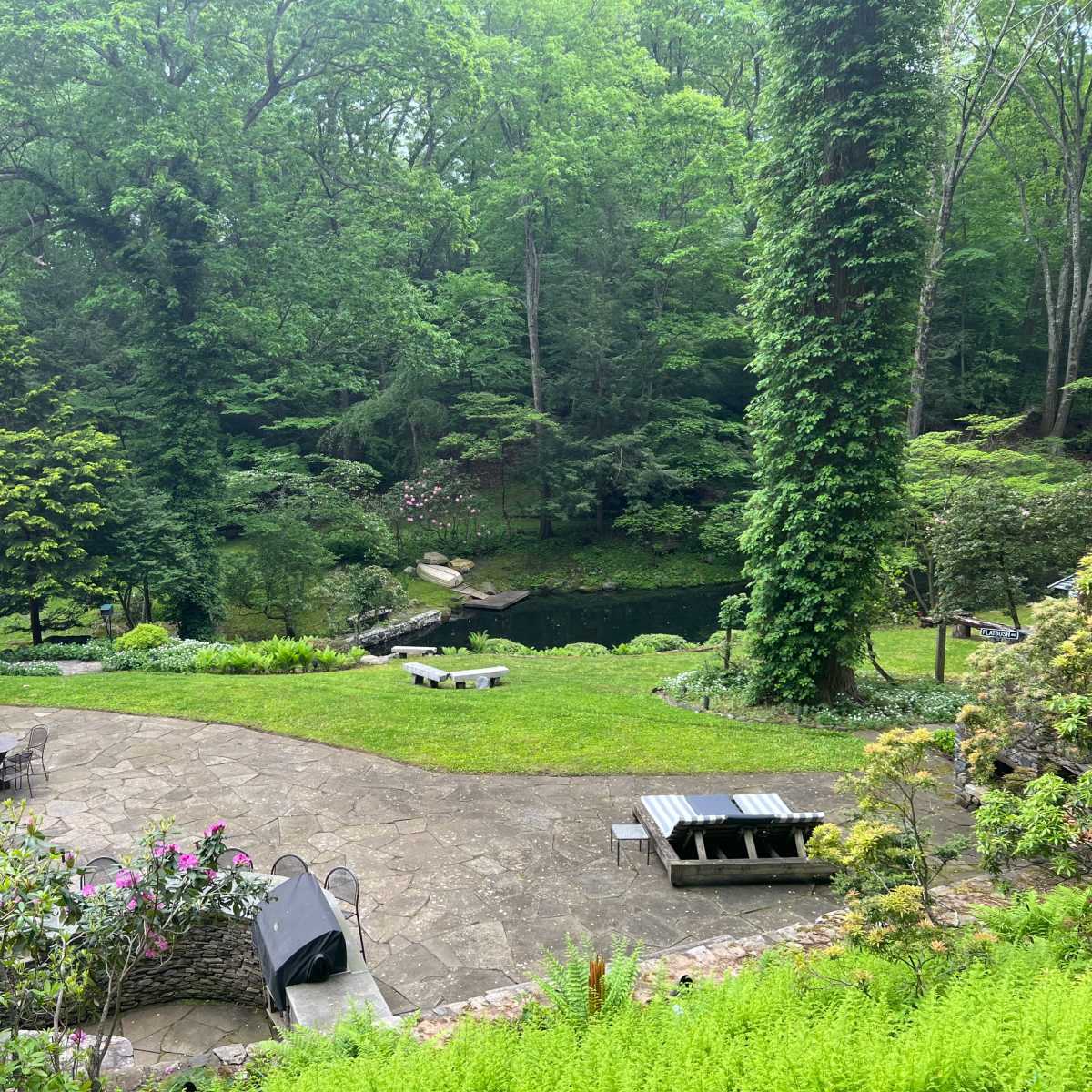 A landscaped backyard with stone pathways, seating areas, and a pond surrounded by lush greenery and flowering plants.