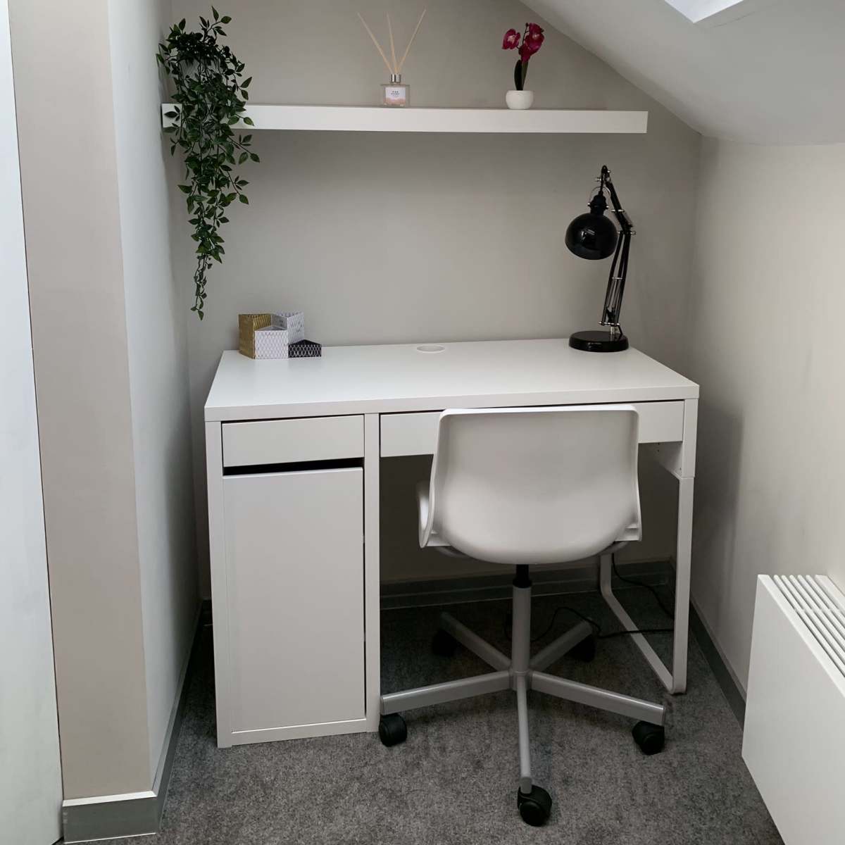 A minimalist desk setup in a corner, featuring a white desk, a swivel chair, a lamp, and decorative items on a shelf above.