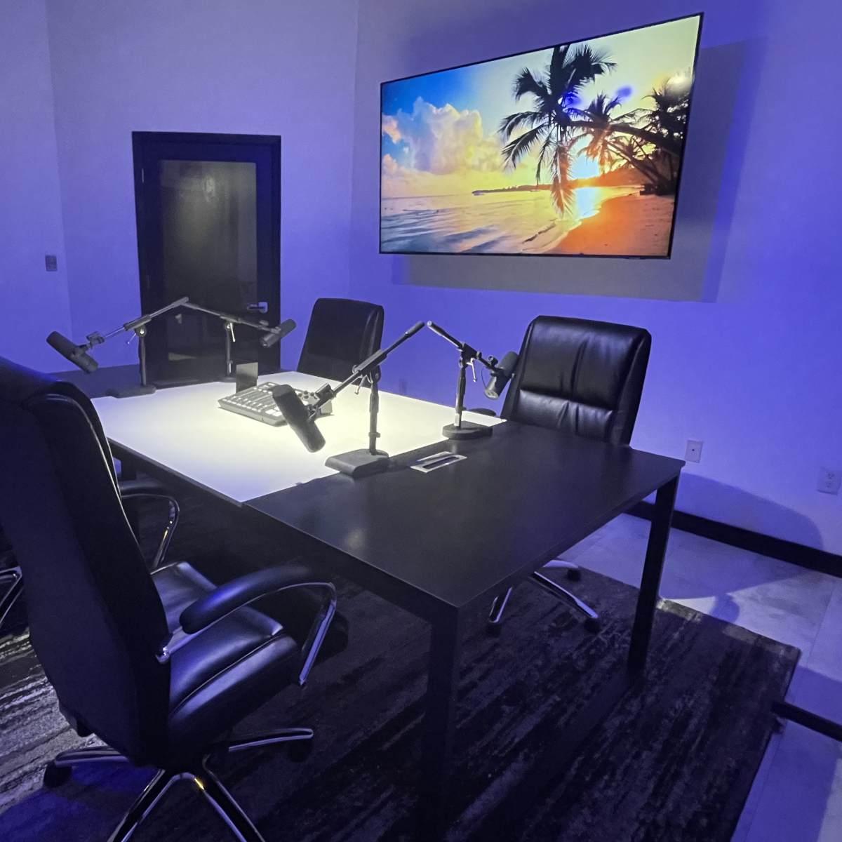 A modern conference room features a large table with four black chairs, illuminated by overhead light, and a large screen displaying a beach scene.