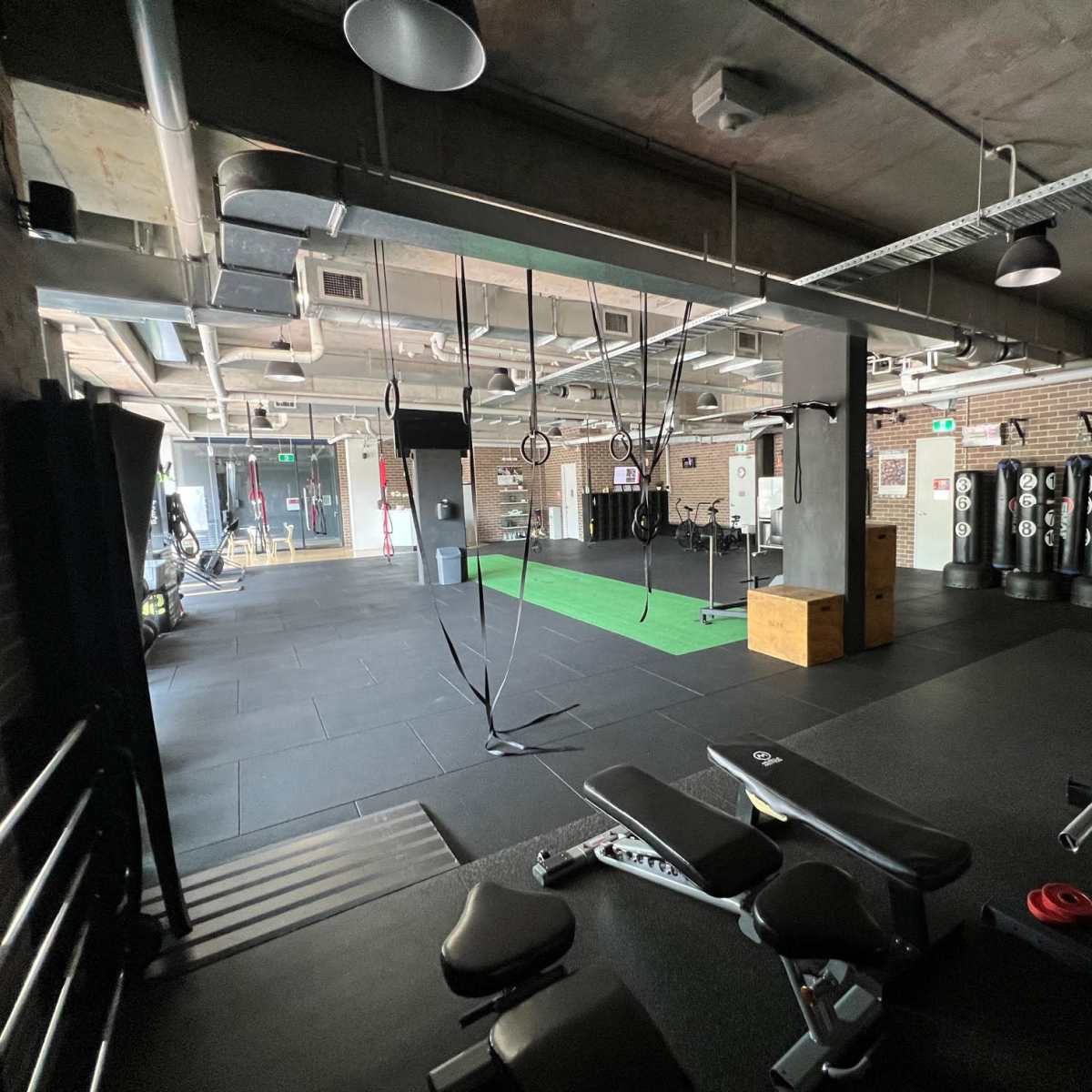 A spacious gym interior with various exercise equipment, including punching bags, a weight bench, and suspended training ropes, all set on black rubber flooring.