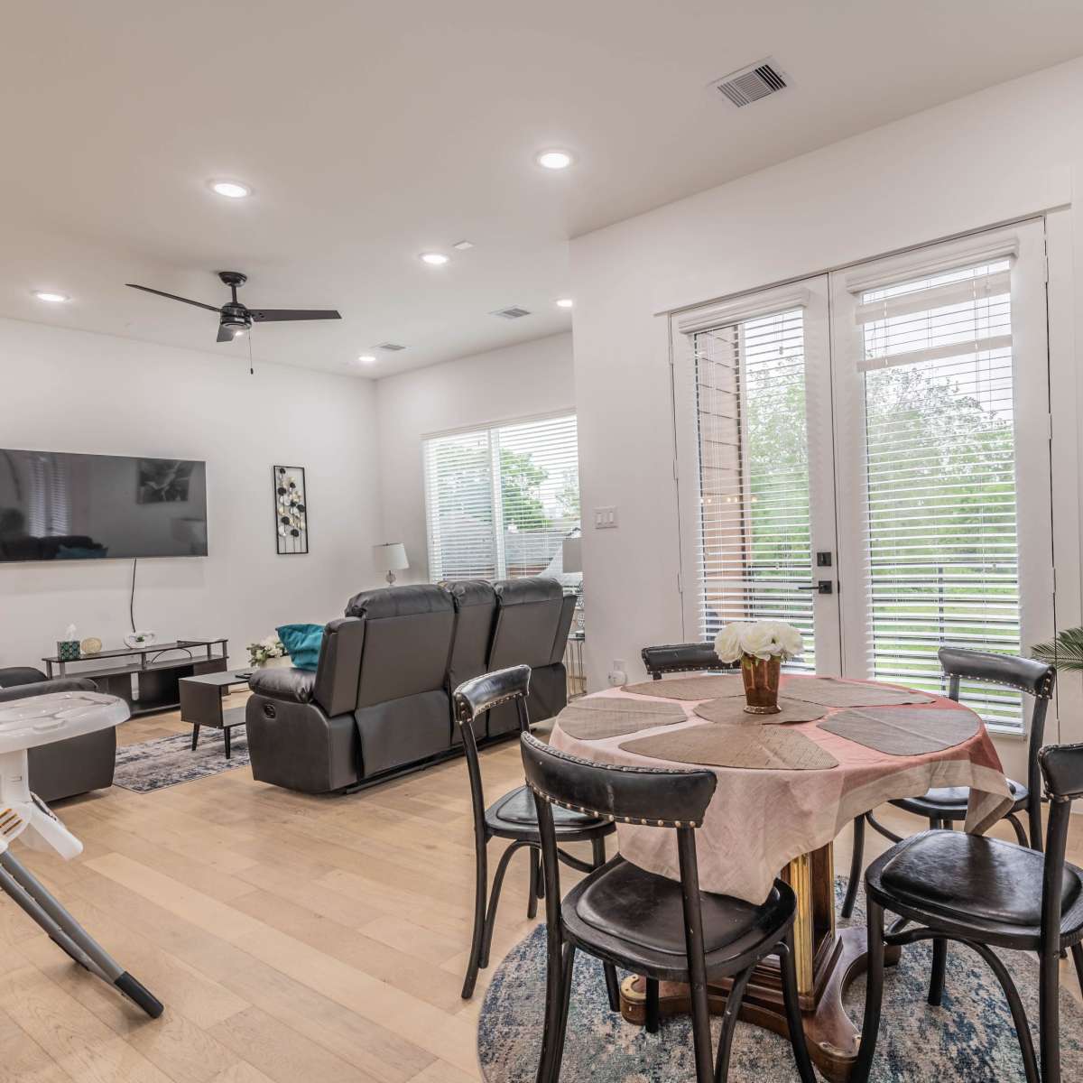 A light-filled living area features a circular dining table with black chairs, a seating area with a television, and large windows that open to a green view.