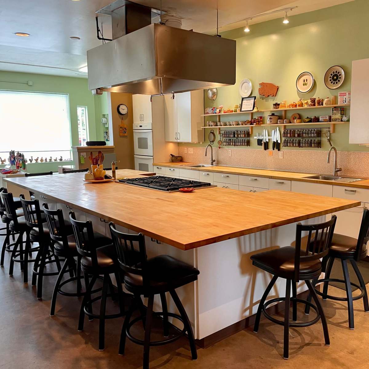 A spacious kitchen with a large central island, equipped with a stovetop, surrounded by black chairs and characterized by white cabinetry and green walls.