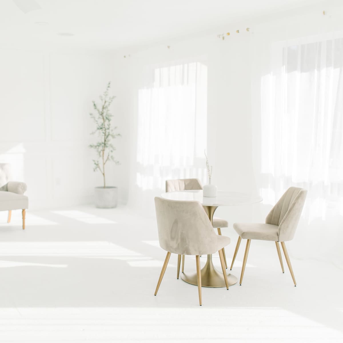 A bright, minimalistic living space featuring a light-colored sofa, two chairs, and a small table, with large windows allowing natural light to flood the room.