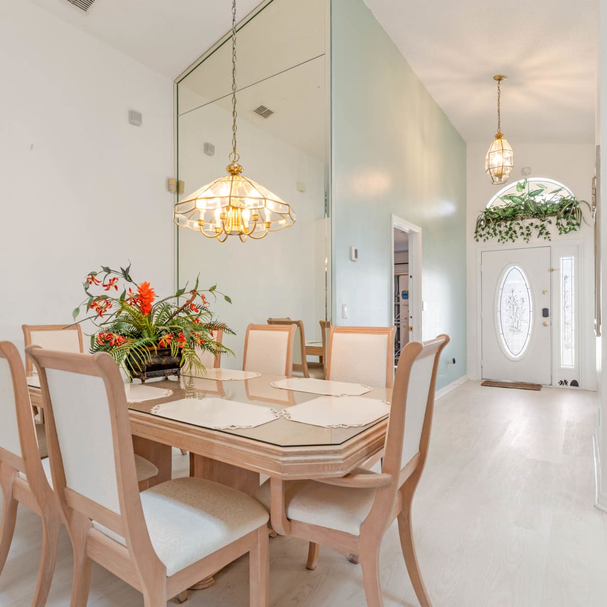 A dining room featuring a wooden table surrounded by eight chairs, with a chandelier overhead and a decorative plant arrangement on the table.
