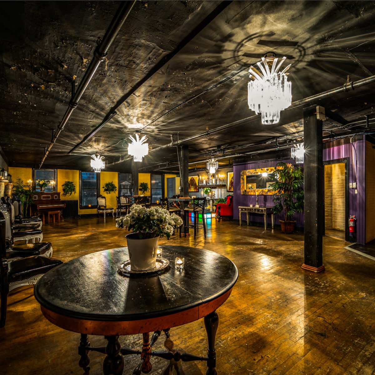 A dimly lit, spacious interior featuring a wooden floor, elegant chandeliers, and various seating arrangements.