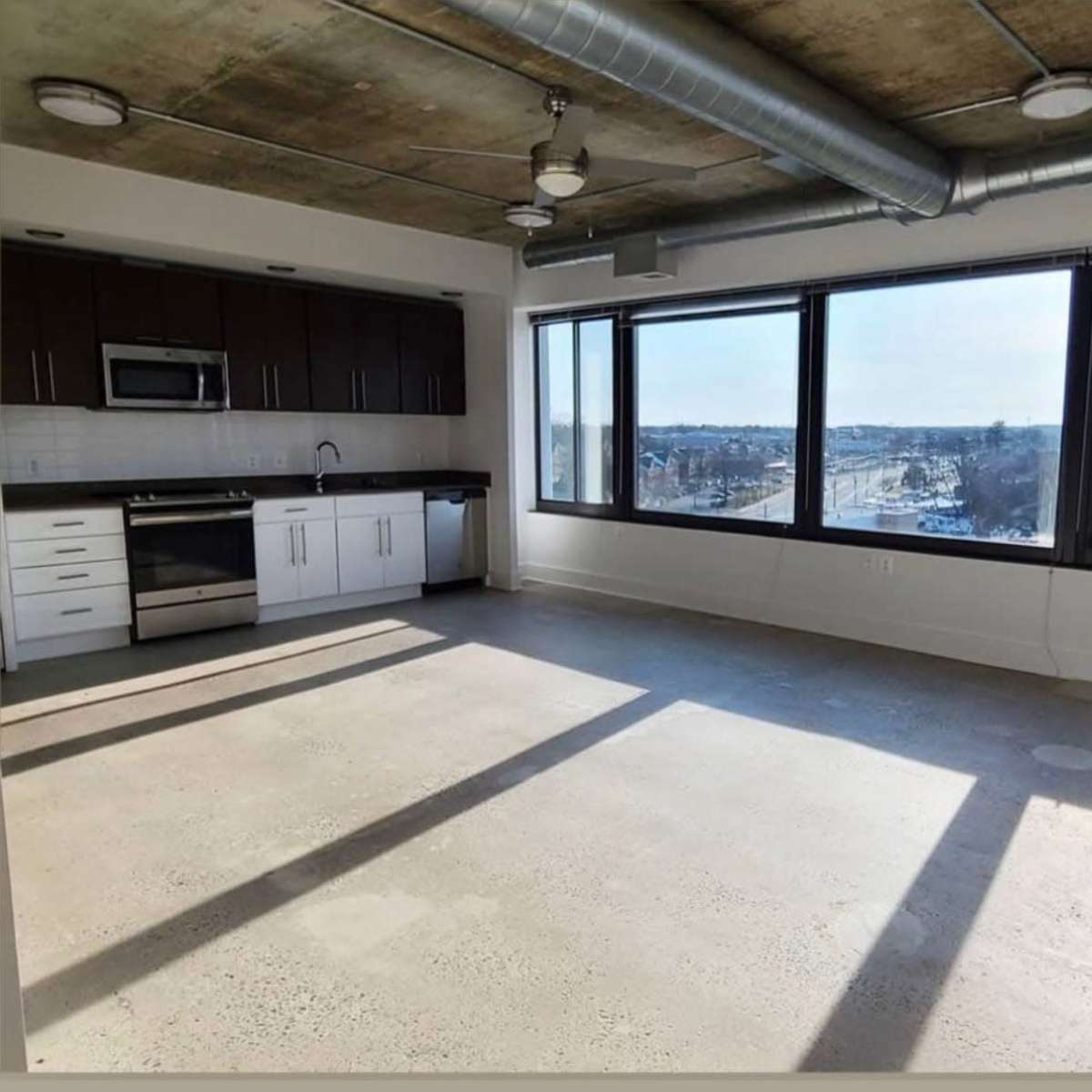 An empty modern apartment with large windows, concrete flooring, and a kitchen area featuring dark cabinetry and stainless steel appliances.