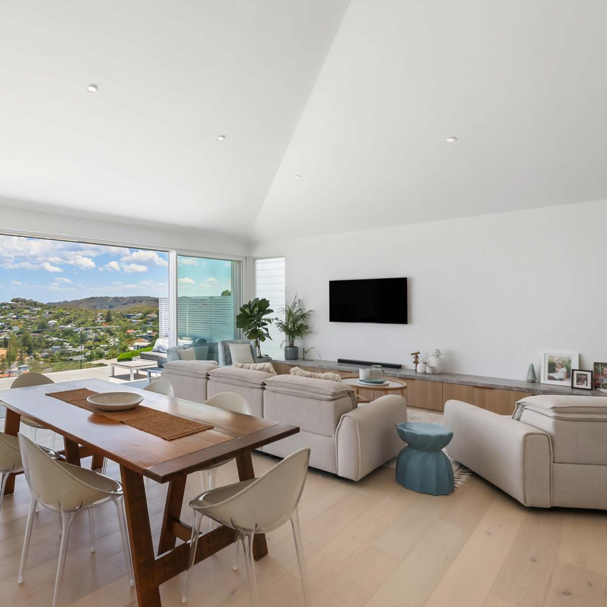 A bright, modern living space features a dining area with a wooden table, surrounded by light chairs, and offers a panoramic view of the beach and coastline through large glass doors.