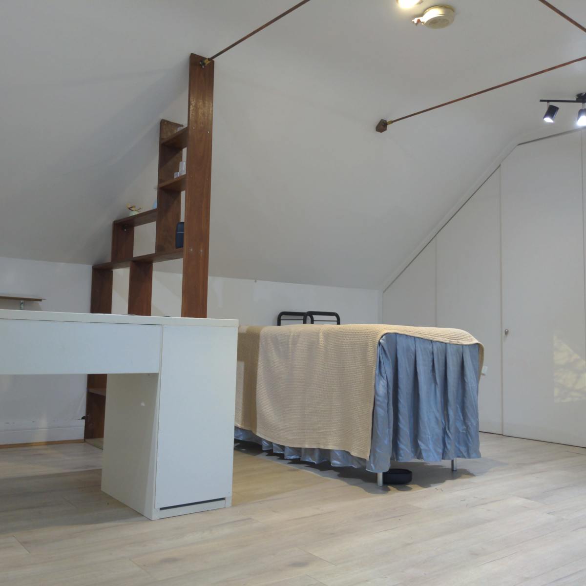 A sparsely furnished attic room featuring a bed with a light-colored bedspread, a desk, and a shelving unit against one wall.