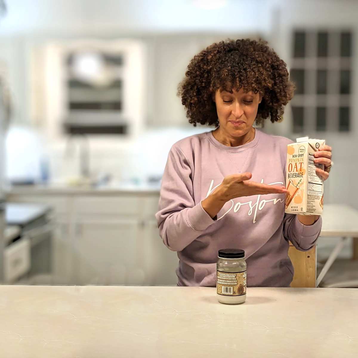 A woman is standing at a kitchen counter, holding a carton of oil in one hand and examining a jar of a different substance in the other.