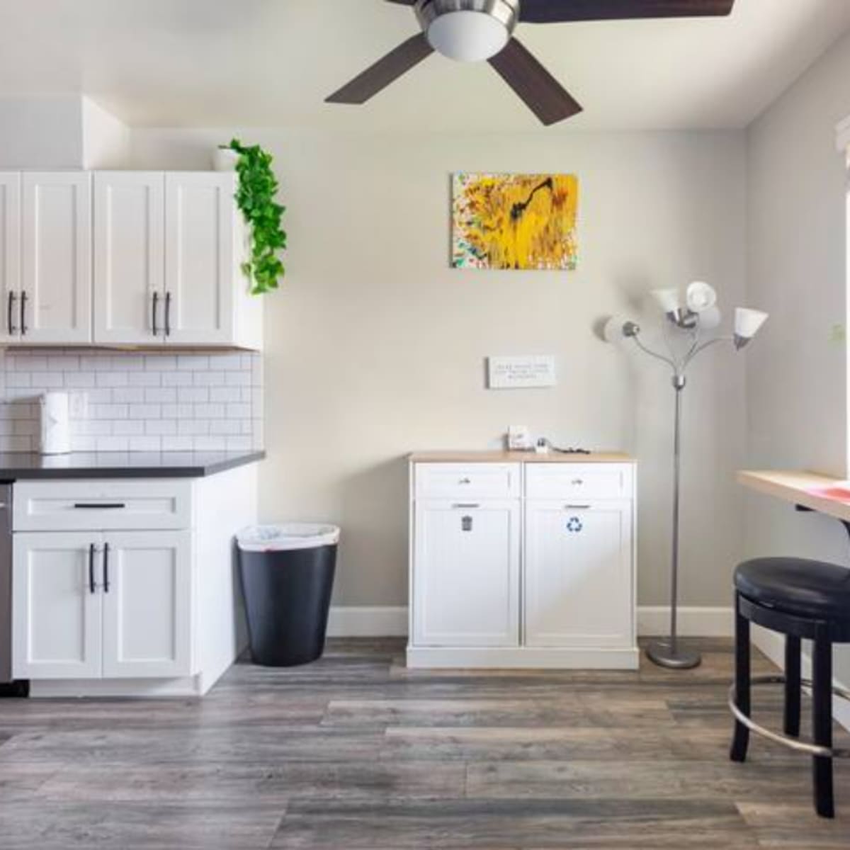 A modern kitchen features white cabinets, a stainless steel refrigerator, a small dining area with two black stools, and a window allowing natural light.