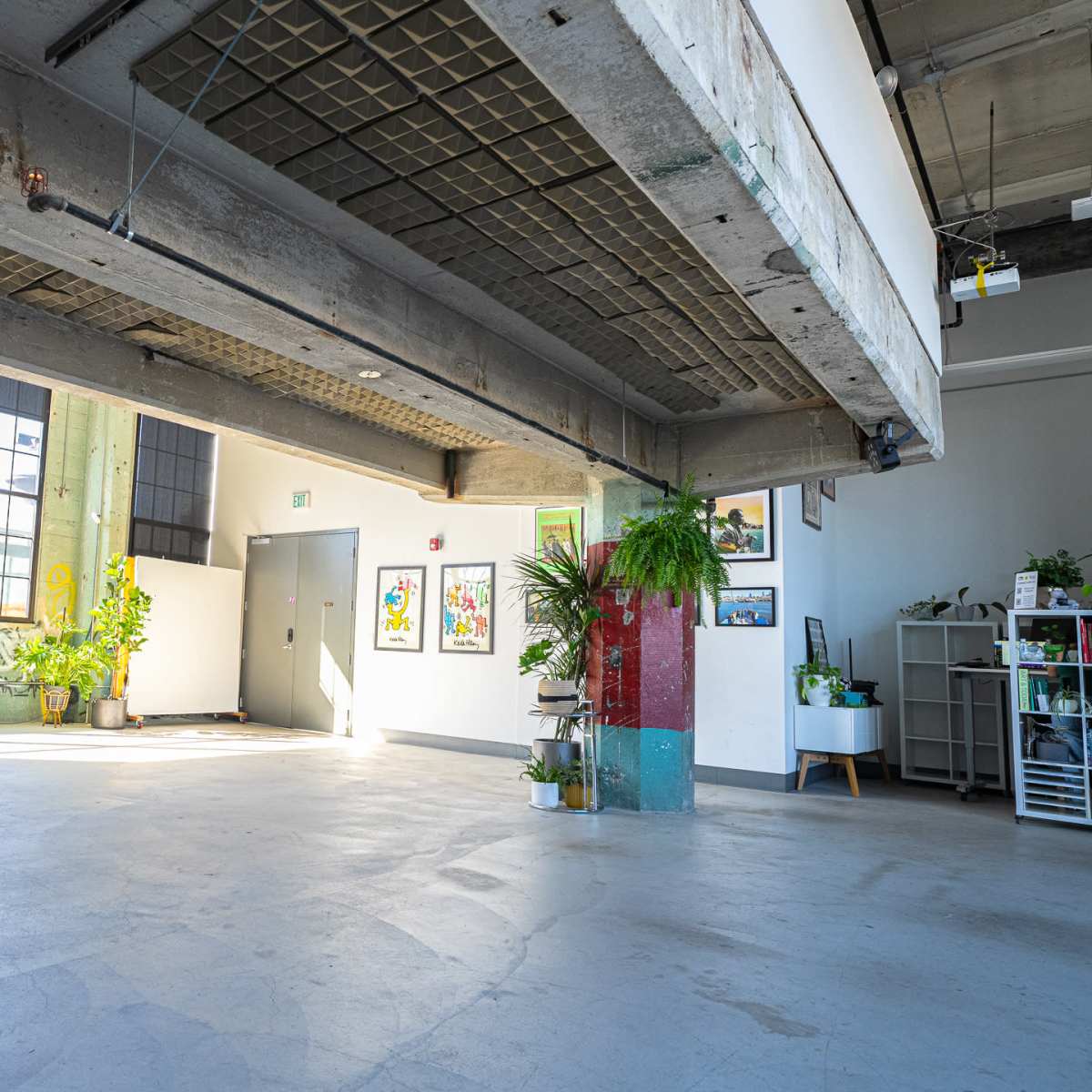 A spacious, industrial-style interior with large windows, exposed concrete beams, and various plants along with artwork on the walls.