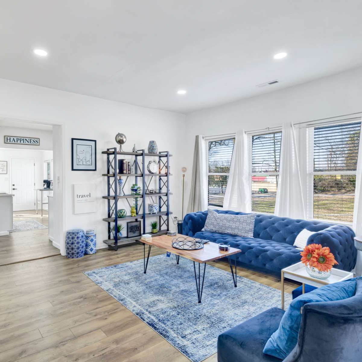 A modern living room features a blue tufted couch, a wooden coffee table, and a bookshelf against a white wall with large windows allowing natural light in.