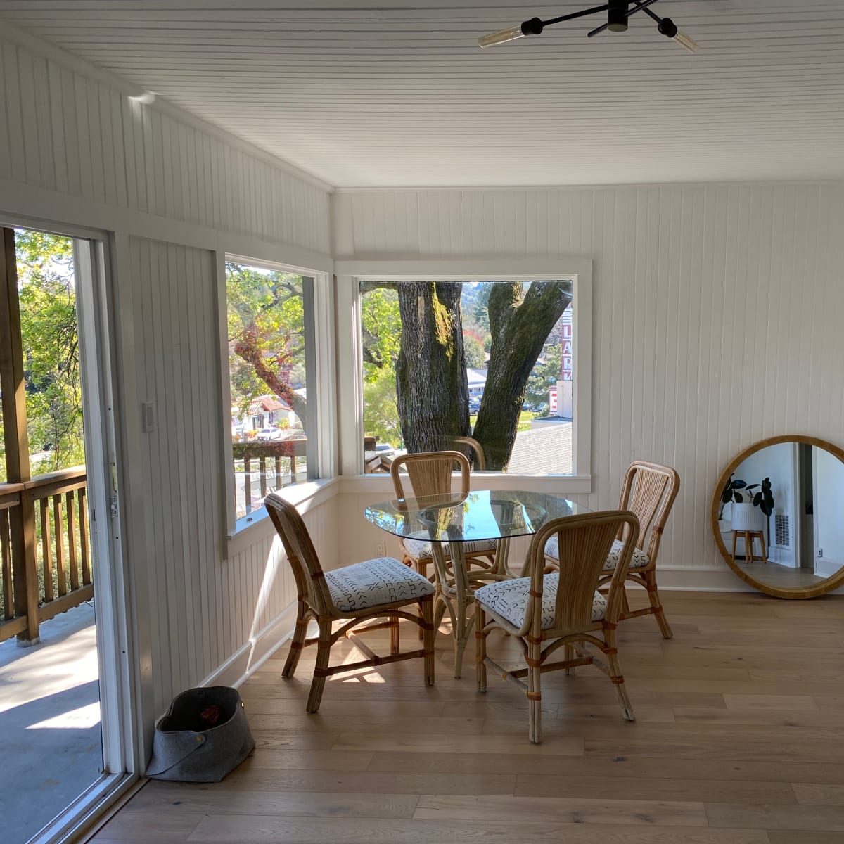 A dining area features a glass table surrounded by four rattan chairs, with a large mirror on the wall and windows overlooking a tree-lined view.