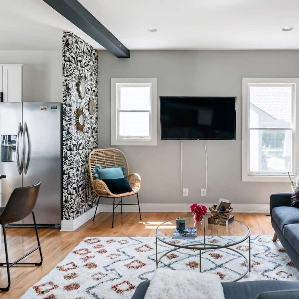 A modern living room features a gray sofa, a glass coffee table, a television mounted on the wall, and a patterned area rug.
