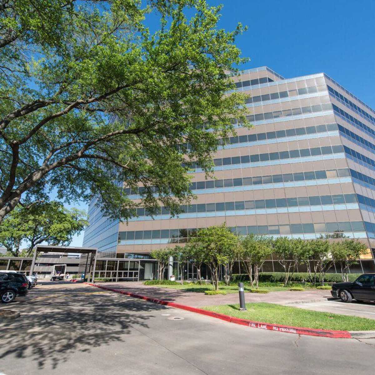 A modern office building with large windows is situated beside a paved parking lot, encircled by trees and landscaping.