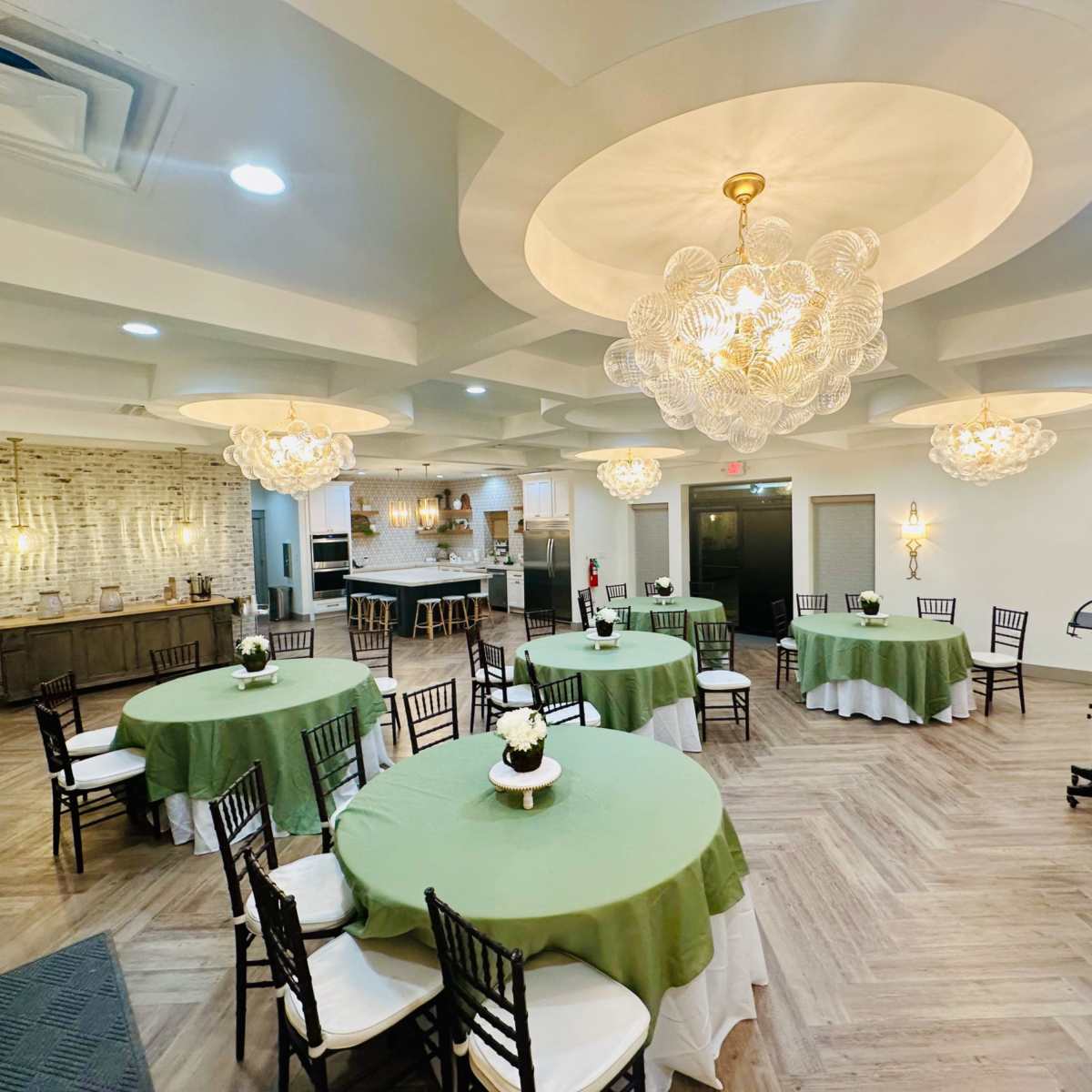 A banquet hall set up with several round tables covered in green tablecloths, each adorned with a small centerpiece, under decorative chandeliers.
