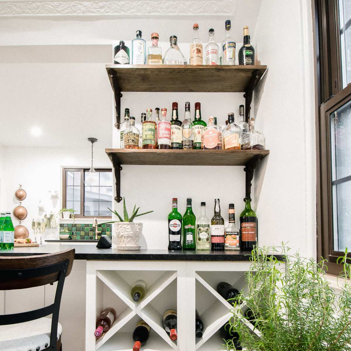 A home bar area featuring a white cabinet with diagonal wine bottle storage, a drawer, a green plant nearby, and shelves above displaying various liquor bottles.