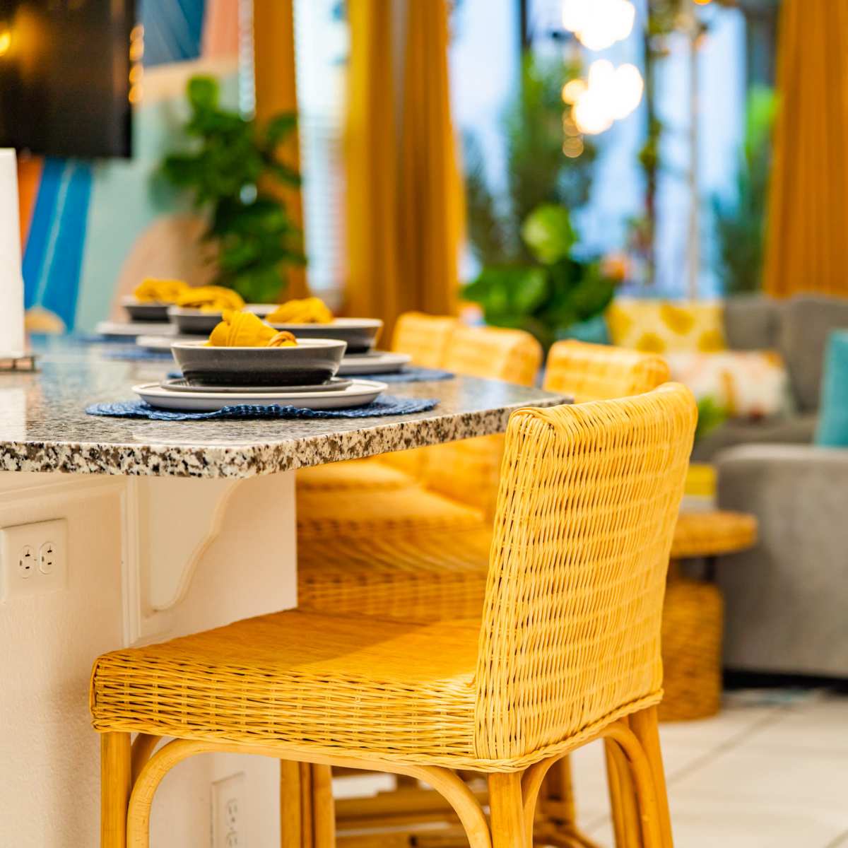 A modern dining area with a granite countertop, wicker bar stools, and a gray sofa in the background.