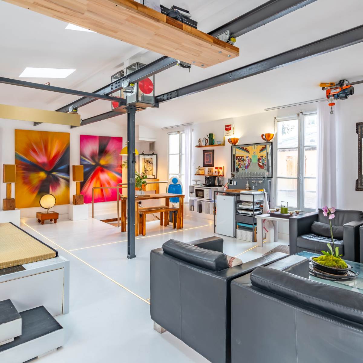A modern interior space featuring a combination of colorful artworks, a seating area with black leather sofas, and various decorative elements, all illuminated by natural light.