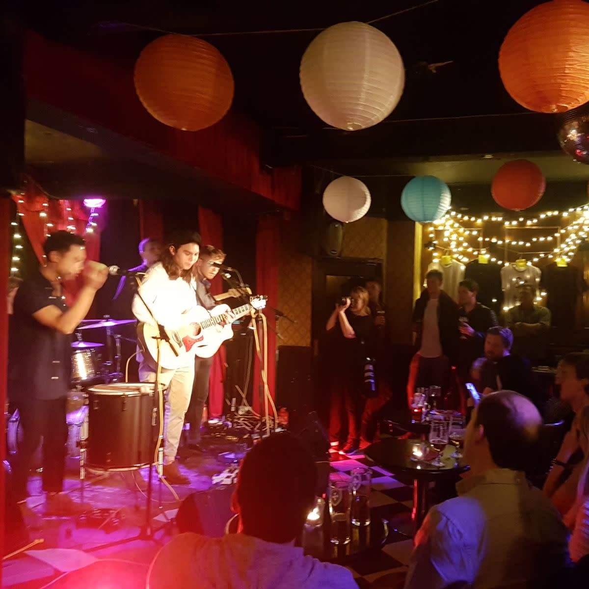 A band performs on stage in a dimly lit bar decorated with colorful lanterns and string lights while an audience watches.
