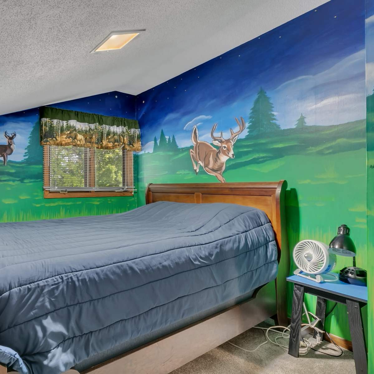 A bedroom with a wooden bed, a painted landscape mural on the walls featuring deer and trees, and a fan beside the bed.