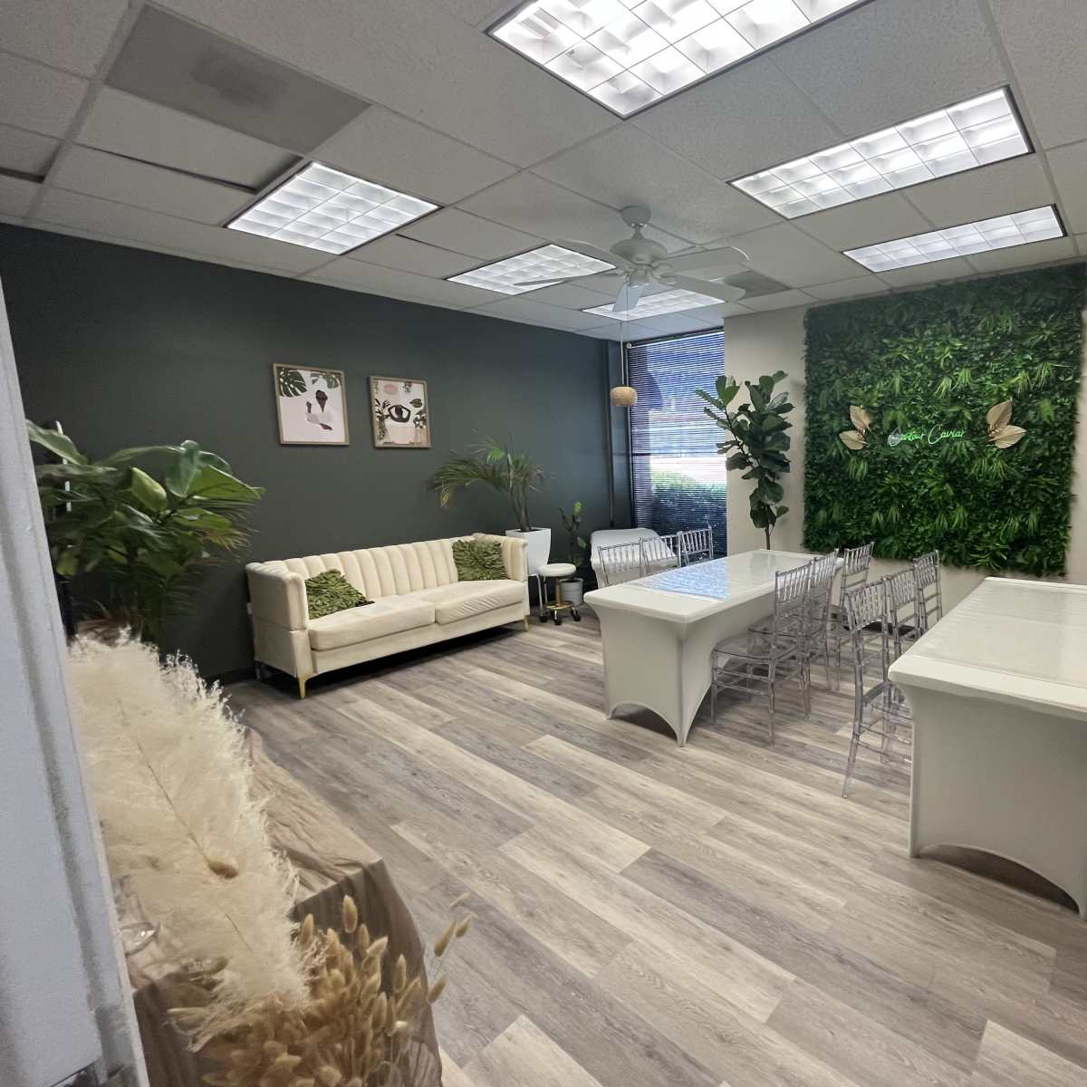 A conference room with a sofa, several transparent chairs, a white table, decorative plants, and a green wall feature.