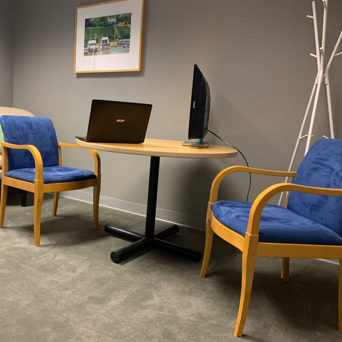 A small office space featuring a round table with a laptop, two blue chairs, and a beige armchair against a gray wall.