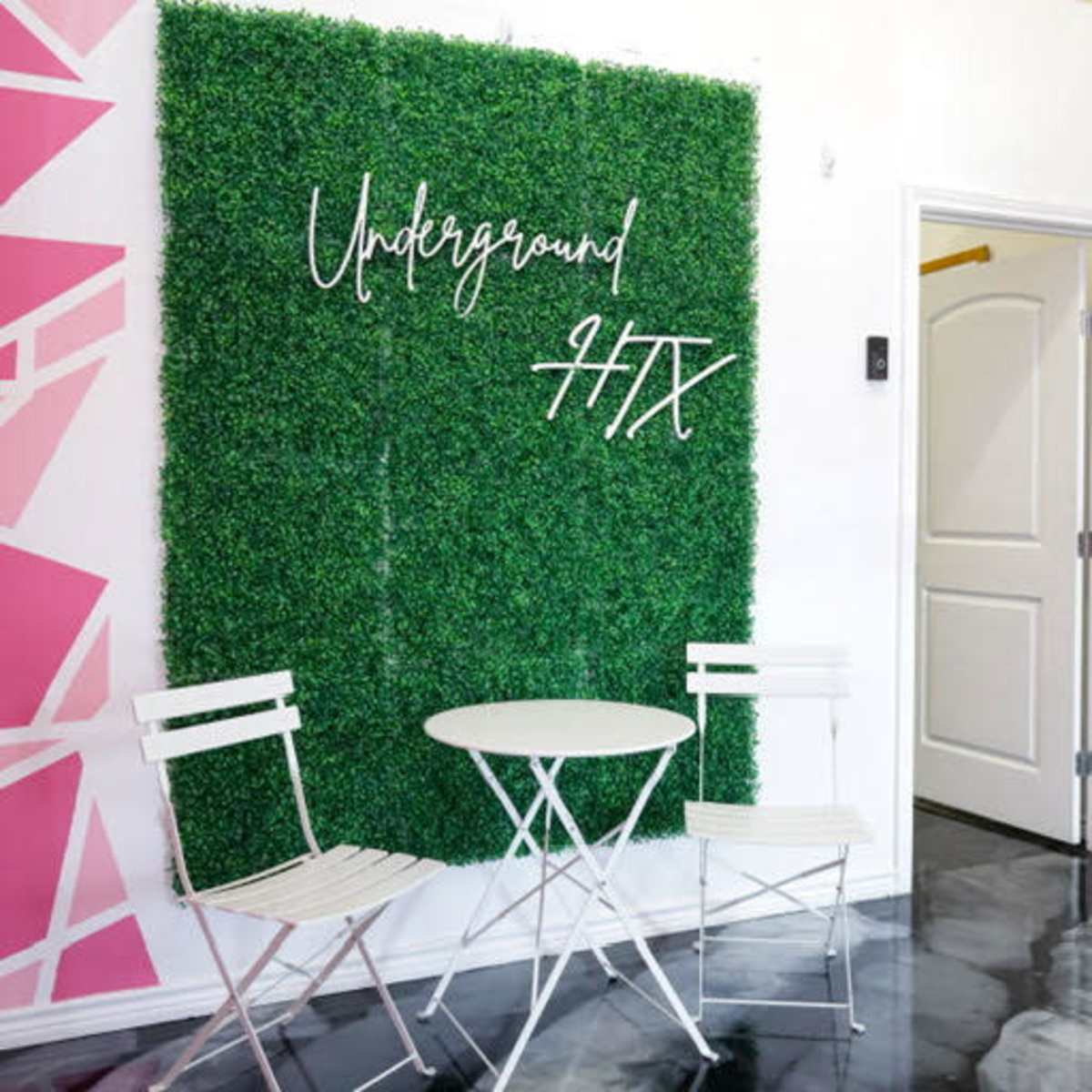 The scene features a small sitting area with a round white table and two white chairs in front of a green artificial grass wall, with the text "Underground HTX" displayed on it.