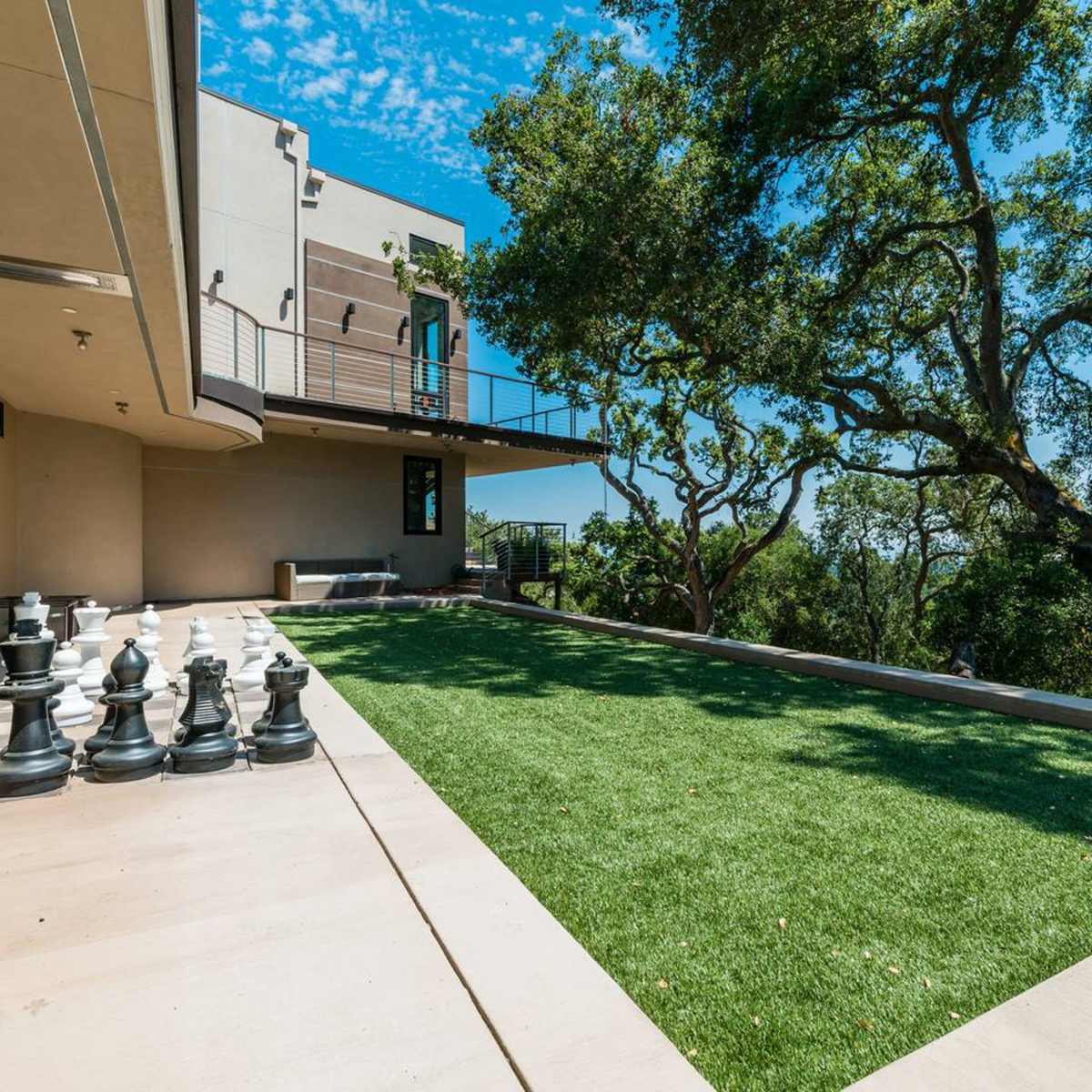 A contemporary outdoor space featuring a large chess set on a paved area next to a green artificial lawn and trees in the background.