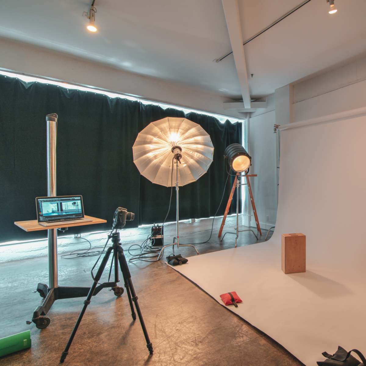 A photography studio setup with a seamless backdrop, a light stand, softboxes, and a tripod positioned near a wooden block.