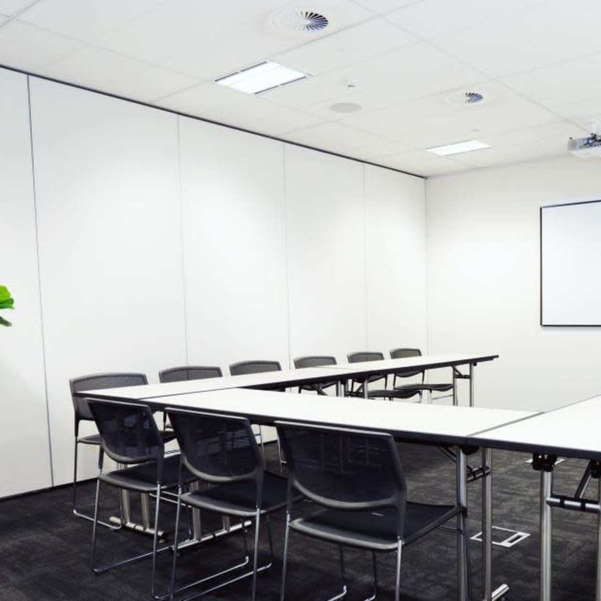 A modern conference room with a long table surrounded by black chairs, two potted plants in the corners, and a whiteboard on the wall.
