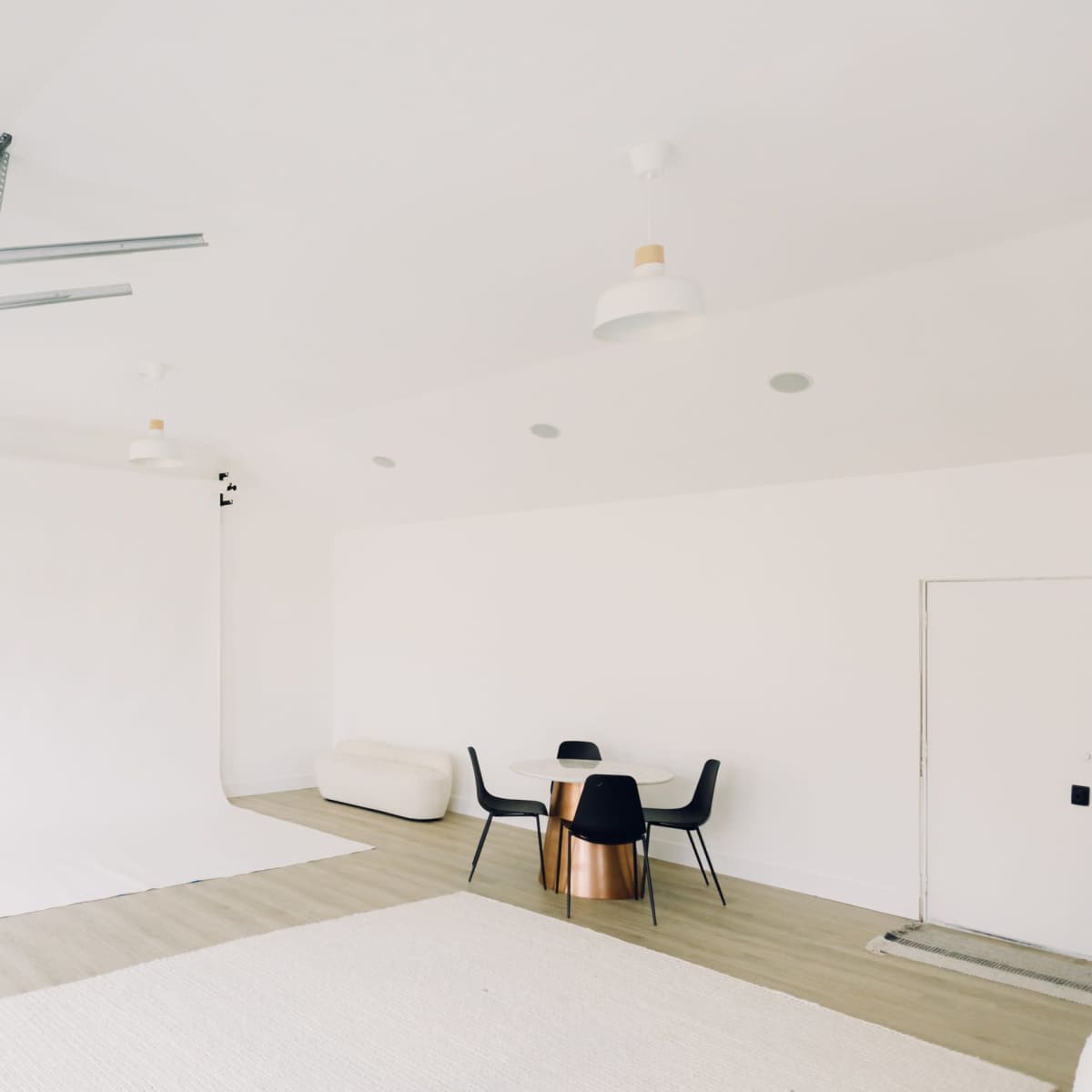 A minimalist studio space with a white backdrop, a round wooden table with black chairs, and a light-colored rug on the floor.