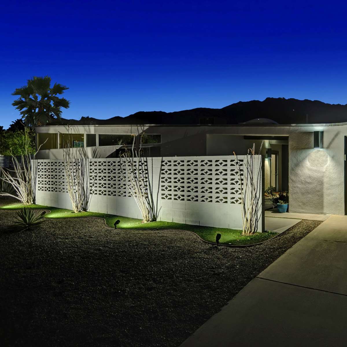 A modern house at dusk, featuring a decorative wall and a garage door, with landscaping illuminated by soft lights.