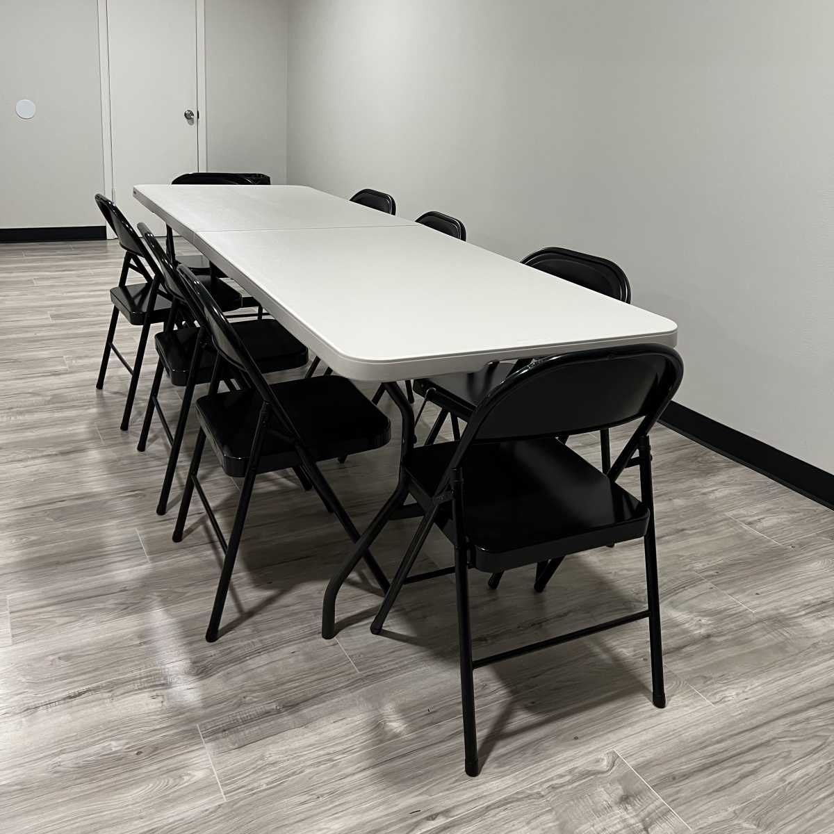 A long, rectangular white table is set in a brightly lit room with six black folding chairs arranged around it.