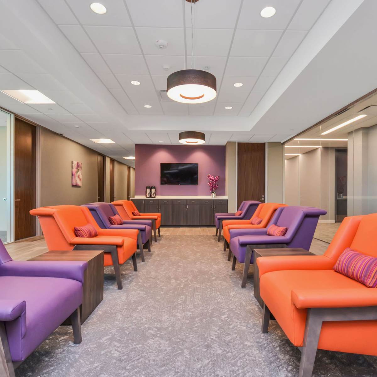 A modern waiting area furnished with purple and orange chairs arranged around a central space, featuring a flat-screen TV and decorative elements on the walls.