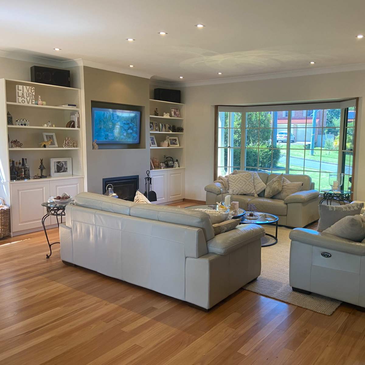 A spacious living room features light-colored leather sofas arranged around a coffee table, with a large window providing natural light and outdoor views.