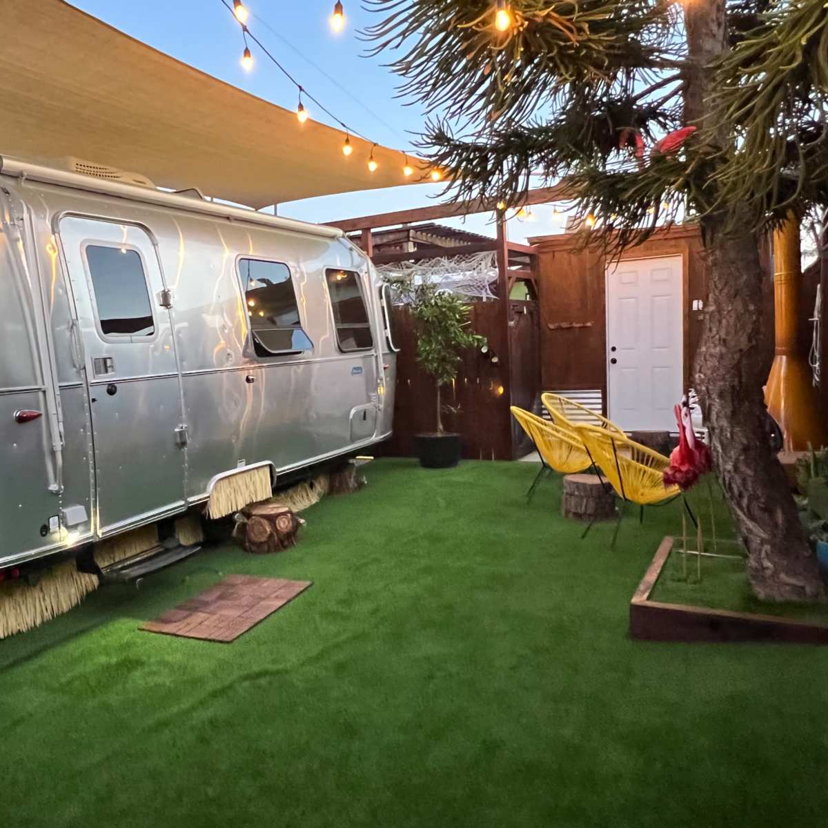 A silver Airstream trailer is parked on a grassy area surrounded by string lights and potted plants.