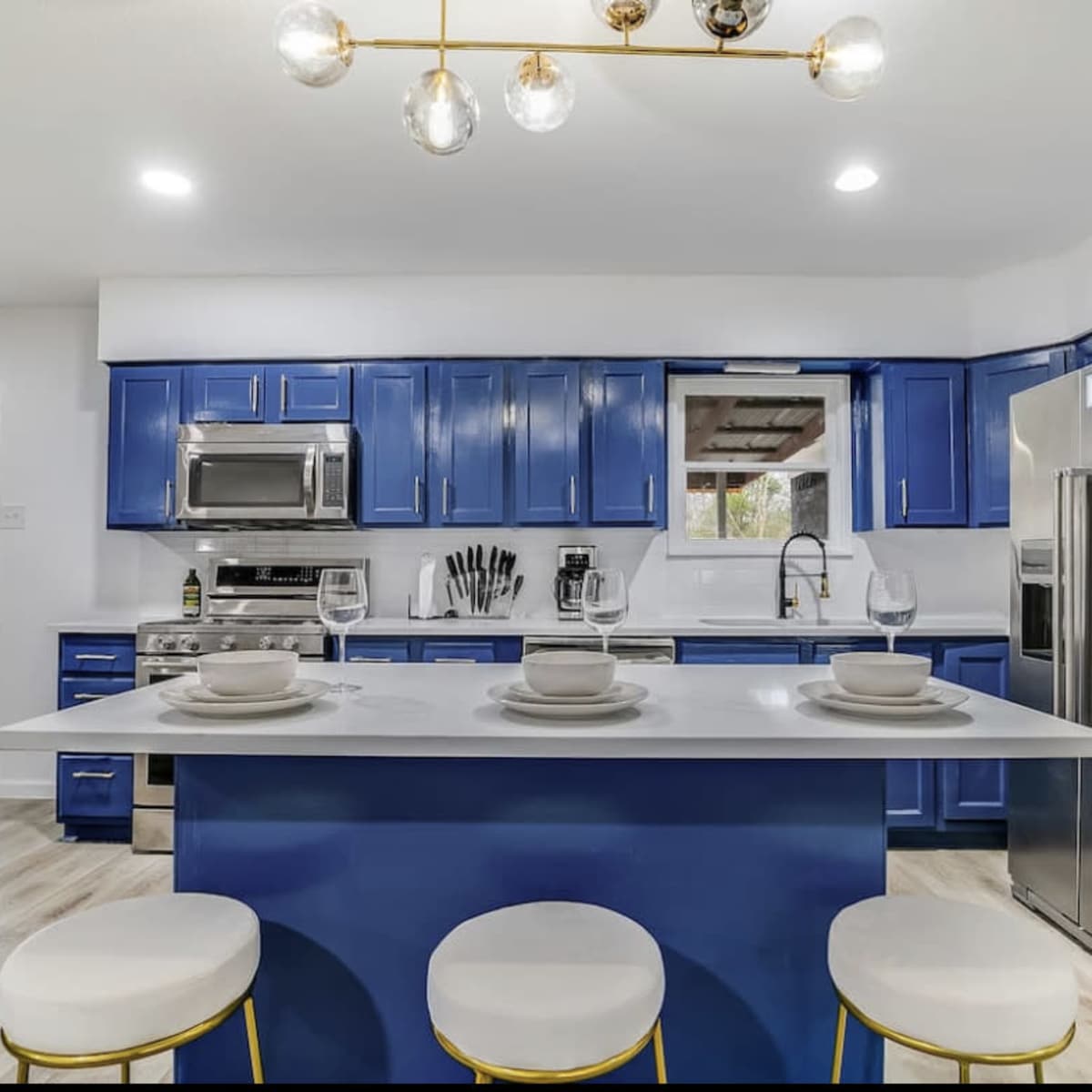 A modern kitchen with blue cabinets, a central island with three white stools, and stainless steel appliances.