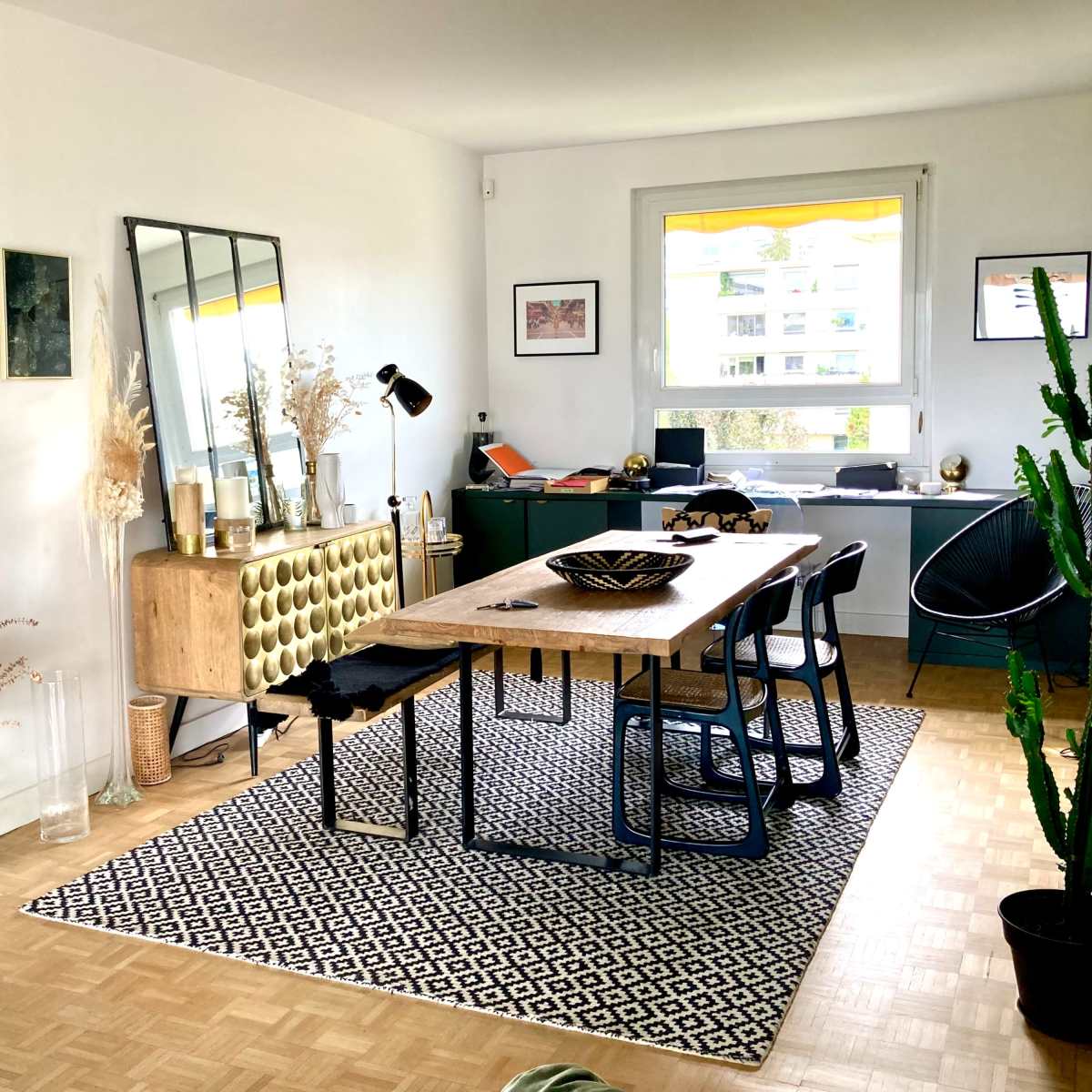 A modern dining area with a wooden table, black chairs, a patterned rug, and decorative plants against a bright, minimalist background.