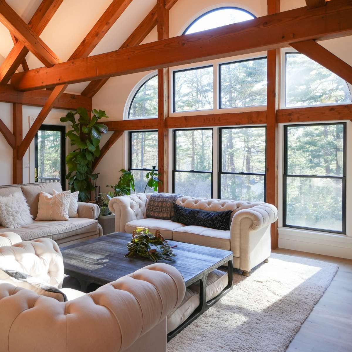 A spacious living room features large windows with wooden beams, showcasing a light-colored sectional sofa and a coffee table on a plush rug.