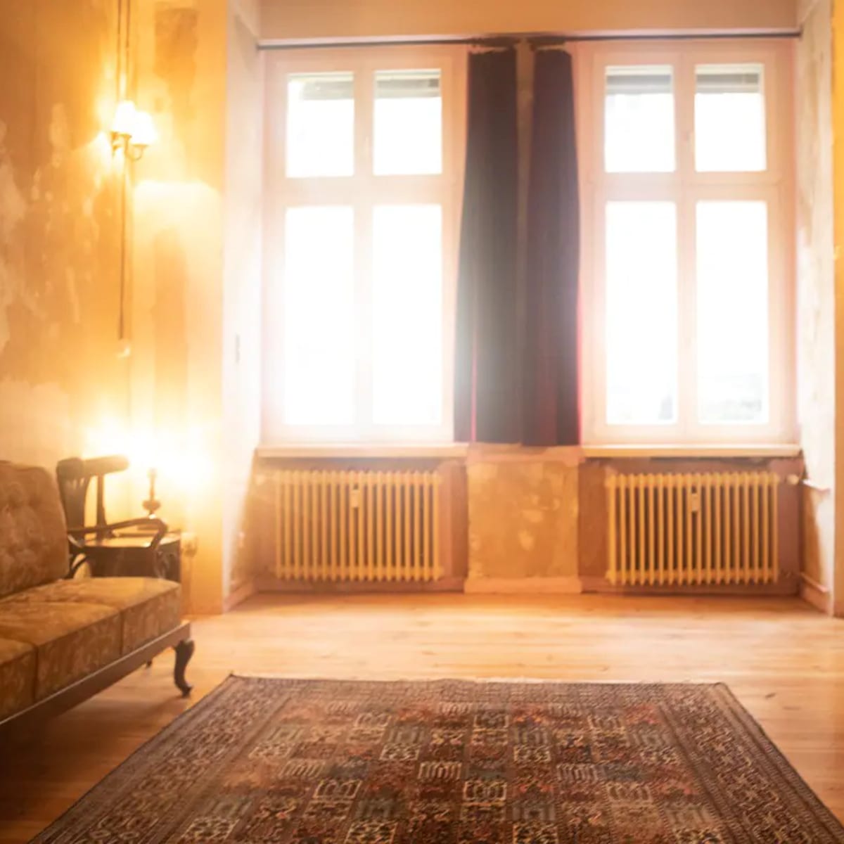 A sparsely furnished room with large windows, exposed walls, a patterned rug, a vintage couch, and an open door.