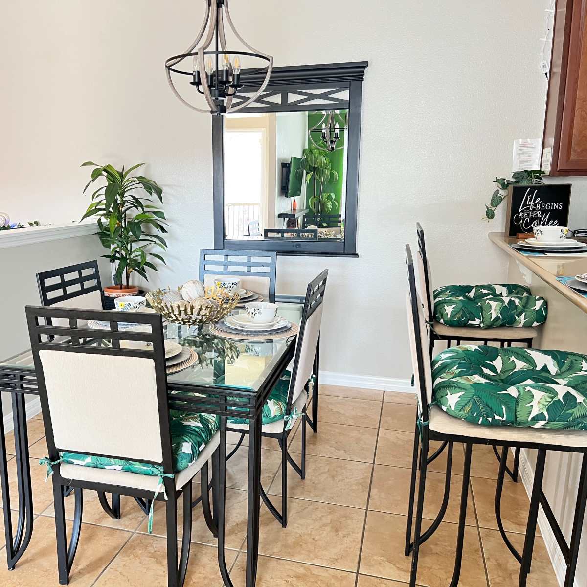 A dining area with a glass-top table surrounded by six chairs, adorned with green tropical-patterned cushions, next to a wall-mounted mirror and a potted plant.