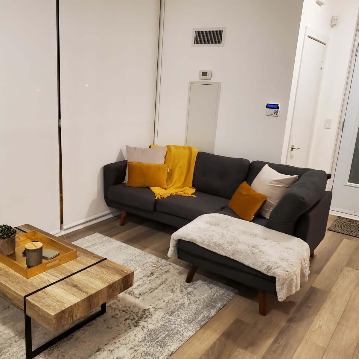 A modern living room features a gray sectional sofa with decorative pillows, a wooden coffee table with candles, and a light rug underfoot.