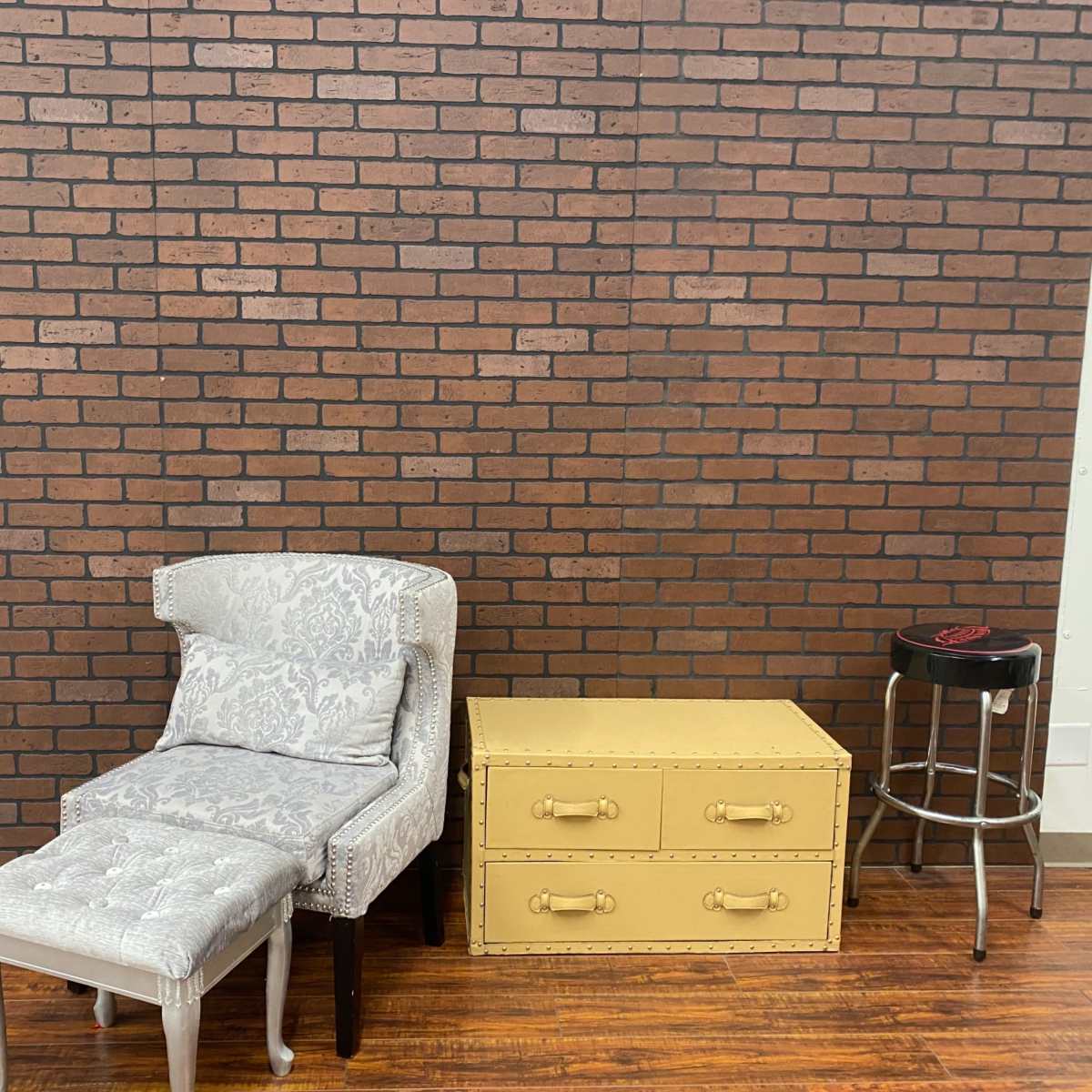 A patterned armchair with an ottoman is positioned next to a yellow storage trunk and a black bar stool against a brick wall.
