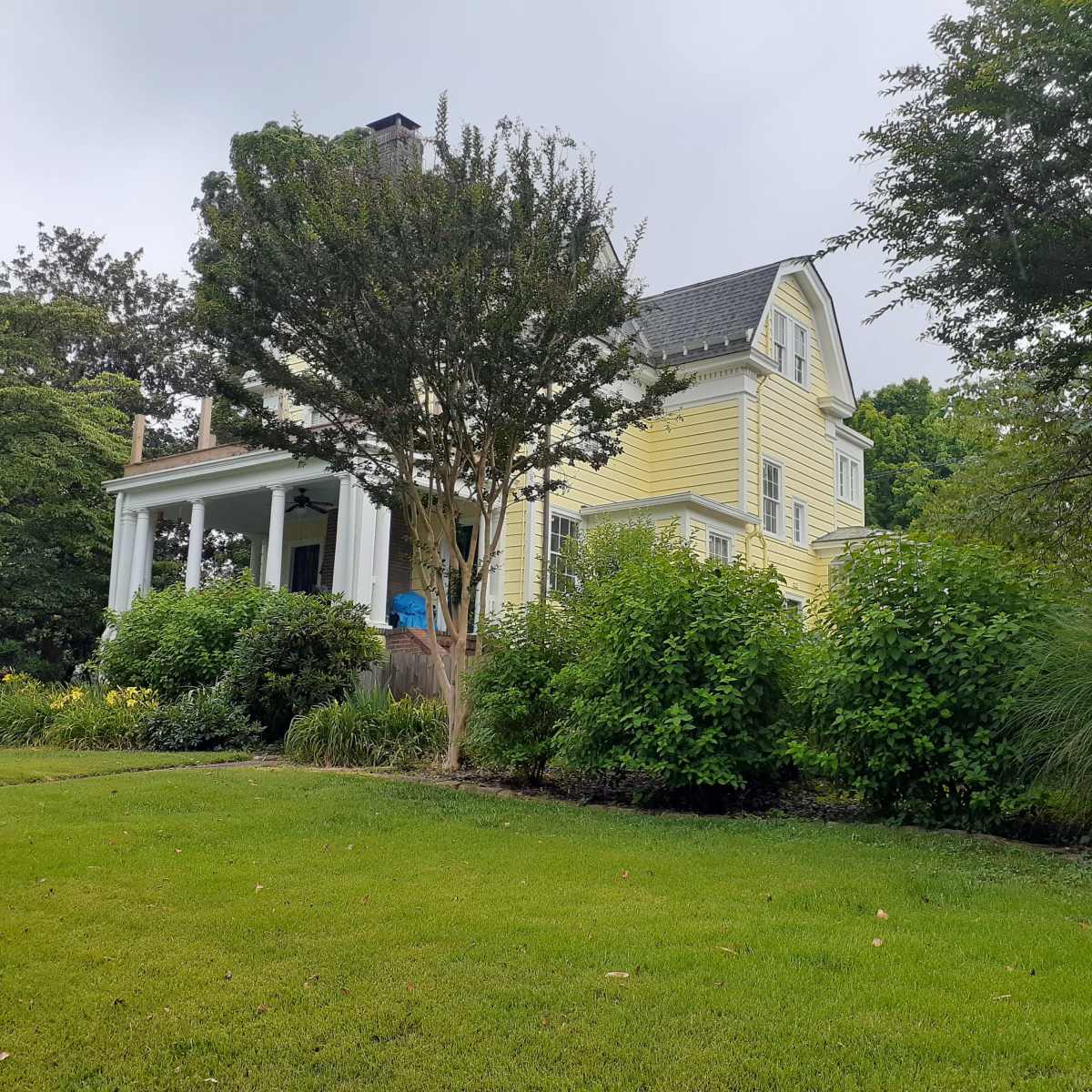 A two-story yellow house with a porch is surrounded by shrubs and trees in a green lawn.