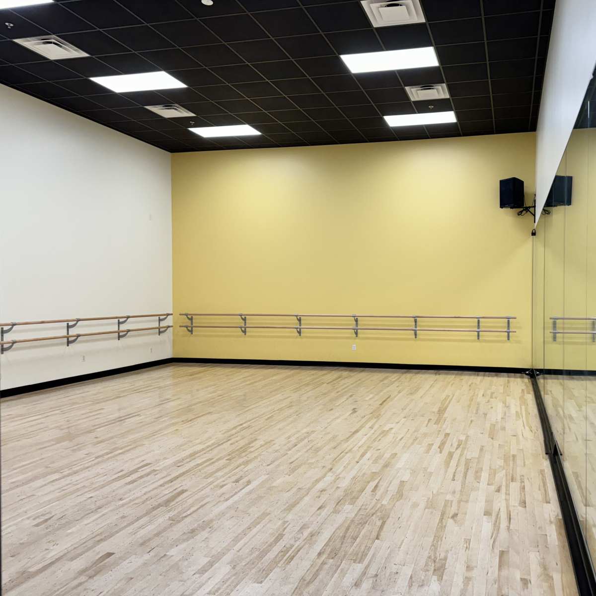 A spacious dance studio with wooden flooring, yellow walls, and a mirrored wall along one side.