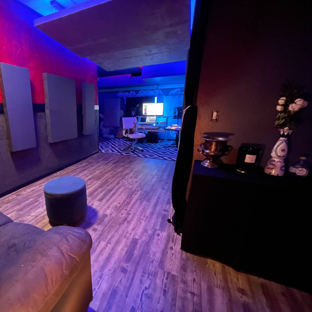 A dimly lit room features a gray couch, a circular ottoman, and a workspace in the background with colorful lighting and musical equipment.